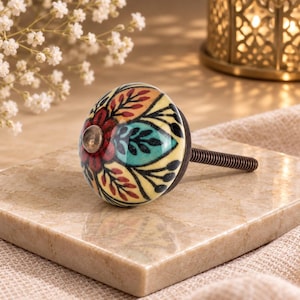 May include: A round ceramic cabinet knob featuring a cream-colored base with a hand-painted floral pattern in shades of red, teal, and black. The knob has a dark metal center accent and a threaded metal bolt extending from the back for installation.