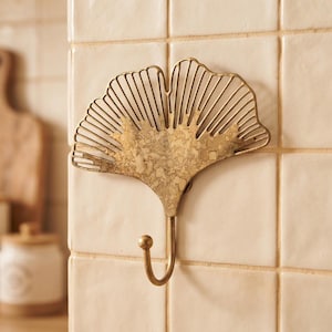 May include: A wall-mounted hook shaped like a ginkgo leaf with a metallic gold finish. The upper portion features a fan-shaped, open-work design with thin vertical slats, while the lower section has a solid, textured surface and a curved hook.