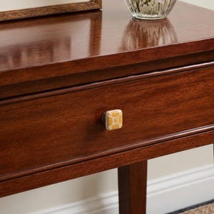 May include: A square cabinet knob with a mustard yellow finish and a white geometric floral pattern centered on a dark brown wooden drawer. The knob features rounded edges and a smooth, glossy surface, contrasting with the rich wood grain texture.