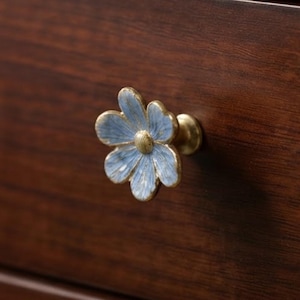 May include: A flower-shaped cabinet knob featuring light blue petals with a distressed finish and a central gold-toned metallic center. The knob is attached to a dark brown wooden surface, showcasing a detailed floral design with visible texture.