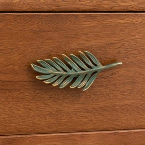 May include: A metal drawer pull shaped like a fern leaf with a weathered green and bronze finish, mounted horizontally on a smooth, medium-brown wooden surface with a visible horizontal grain pattern throughout the wood texture.