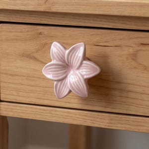 May include: A light pink ceramic drawer knob shaped like a six-petaled flower with embossed vertical lines on each petal. The knob is attached to a light-toned wooden drawer front with a visible horizontal wood grain pattern and smooth finish.
