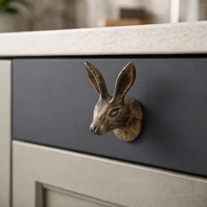 May include: A bronze-toned metal cabinet knob shaped like a rabbit head with long, upright ears and detailed facial features. The knob is mounted onto a dark gray drawer front, providing a functional handle for opening and closing the cabinet.