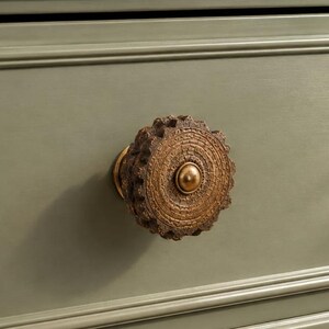 May include: A round, bronze-colored drawer knob with intricate, concentric circular carvings and a smooth, raised center dome. The knob is mounted on a flat, sage green surface featuring horizontal molding lines across the top and bottom edges.