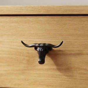 May include: A matte black cabinet knob shaped like a bull head with long, curved horns. The hardware is mounted onto a light-colored, natural wood drawer front with a visible horizontal grain pattern, creating a contrast between the dark metal and wood.