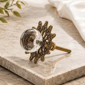 May include: A decorative cabinet knob featuring a clear, spherical glass center mounted on an ornate, sunburst-shaped antique brass metal backplate with a threaded metal bolt extending from the rear for installation purposes.