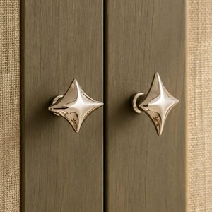 May include: Two polished silver-toned metal cabinet knobs with a four-pointed star shape. Each knob features a smooth, reflective surface and a central point that tapers outward to four distinct corners, mounted on a vertical wooden cabinet door.