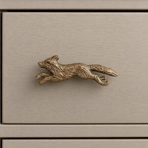 May include: A brass-toned metal drawer knob shaped like a running fox. The fox figure features a textured, weathered finish with visible details of its fur, ears, and tail, positioned horizontally as a functional handle on a flat surface.
