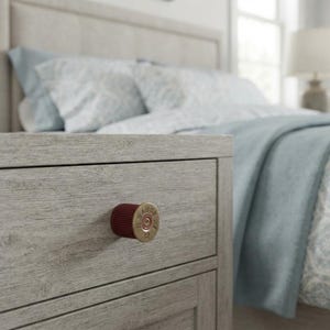 May include: A light gray wood grain nightstand drawer featuring a single drawer pull made from a maroon and brass 12 gauge shotgun shell casing. The brass base of the shell is clearly visible, displaying the text 12 GAUGE and other markings.