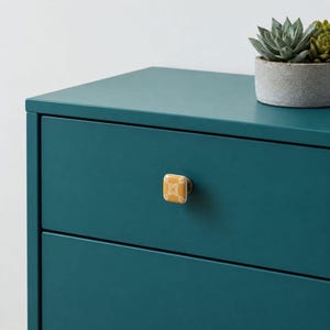 May include: A square mustard yellow ceramic cabinet knob with a white geometric floral pattern attached to a teal drawer. The knob features a metallic base and is mounted on a solid teal furniture piece with a succulent in a gray pot above.