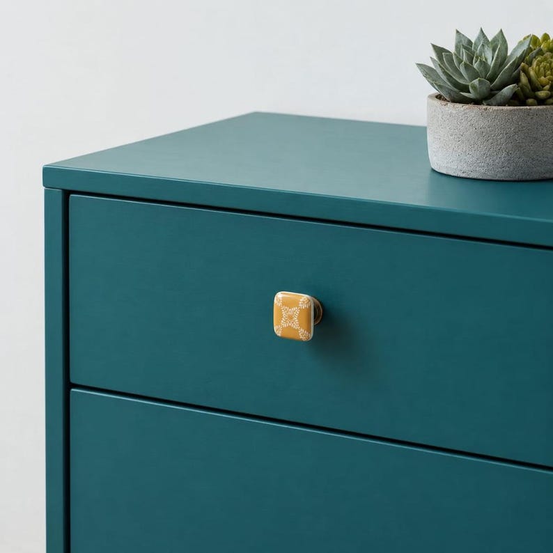 May include: A square mustard yellow ceramic cabinet knob with a white geometric floral pattern attached to a teal drawer. The knob features a metallic base and is mounted on a solid teal furniture piece with a succulent in a gray pot above.