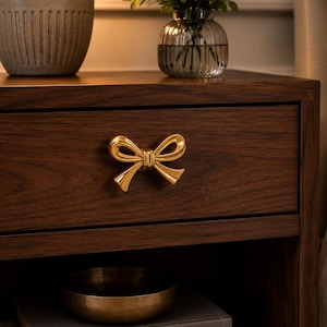 May include: A dark brown wooden nightstand featuring a single drawer with a polished gold-toned metal bow-shaped handle. The drawer front displays a prominent wood grain pattern, and the hardware is centered on the drawer face.