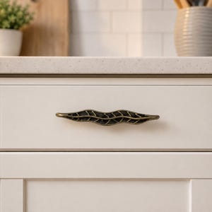 May include: A horizontal cabinet pull with an elongated, wavy leaf-like shape. The hardware is finished in a dark, antique brass tone with a textured, open-work design that features a central vein and symmetrical side segments.