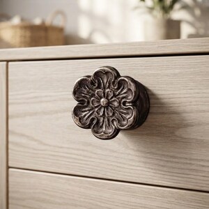 May include: A dark metallic drawer knob shaped like a six-petaled flower with an antiqued finish. The knob is mounted on a light-colored wooden drawer front with a visible horizontal wood grain pattern, featuring a central circular detail.