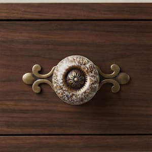 May include: A round ceramic drawer knob with a speckled brown and cream finish, featuring a central bronze-toned floral metal inlay. The knob is mounted on a decorative, symmetrical bronze-toned metal backplate with curved, scroll-like extensions.