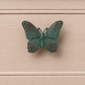 May include: A butterfly-shaped drawer knob with a dark green, weathered patina finish. The metal hardware features intricate embossed wing patterns with raised circular details and a central body, mounted on a light beige wood surface.