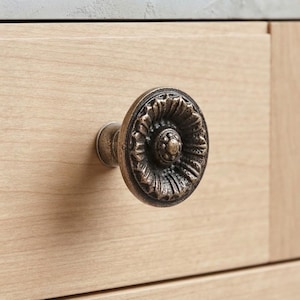 May include: A round, bronze-toned metal cabinet knob with a textured, floral-inspired relief pattern. The knob features a central circular element surrounded by radiating petals, mounted on a light-colored wooden drawer front with a smooth finish.