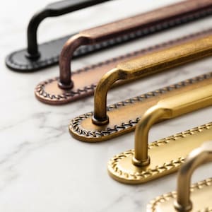 May include: A series of metal cabinet pulls arranged in a diagonal row. Each handle features a long, slender bar attached to an oval-shaped backplate with a decorative braided or rope-like border. The finishes include matte black, copper, bronze, and gold.