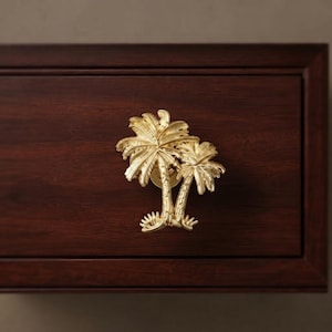 May include: A gold-toned metal cabinet knob shaped like two palm trees with textured trunks and fronds. The hardware is mounted onto a dark brown wooden drawer front with a visible grain pattern, featuring a rectangular frame design.
