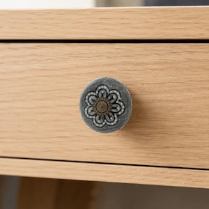 May include: A round cabinet knob featuring a dark gray surface with a white floral pattern and a central bronze-toned metal accent. The knob is attached to a light-colored wooden drawer front with a visible horizontal wood grain texture.