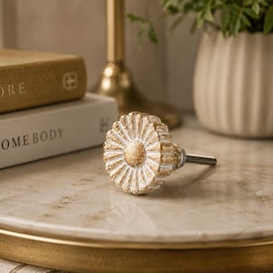 May include: A round drawer knob featuring a flower-like design with radiating petals. The surface has a distressed finish, combining metallic gold accents with white paint. A threaded metal bolt extends from the back of the circular knob.