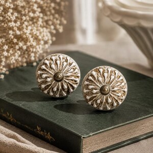 May include: Two round cabinet knobs featuring a raised white floral petal pattern over a distressed brown base. Each knob has a central brass-toned metal stud. The design consists of radiating white lines that extend from the center to the edge.