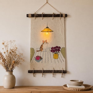 Op de afbeelding: Een rechthoekige stoffen wandhanger opgehangen aan twee donkere houten takken en touw. De illustratie toont een kat op een tafel met een roze mok, een gele vaas met rode rozen en een oplichtende gele hanglamp die erboven hangt.