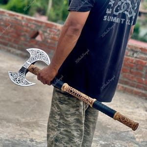 May include: A double-bitted battle axe featuring a silver-colored head with intricate black Celtic knotwork engravings. The long wooden handle is carved with matching patterns and includes a black wrapped grip section near the base of the shaft.