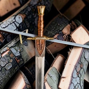 Può includere: Un pugnale metallico con impugnatura color rame a coste, pomolo e guardia color oro. Il fodero è decorato con un emblema a scudo centrale contenente un simbolo di tridente e motivi a chevron incisi lungo la lama.