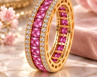 Op de afbeelding: Een gouden eeuwigheidsring met een centrale rij vierkant geslepen roze edelstenen, geflankeerd door twee rijen kleine, ronde, helderwitte stenen. De ring heeft een gepolijste gouden afwerking en een doorlopend kanaalontwerp rondom.