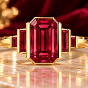 May include: A gold ring featuring a large emerald-cut red gemstone in the center, flanked by two smaller emerald-cut red gemstones on each side. The stones are set in a polished yellow gold band with a clean, structured metal frame.
