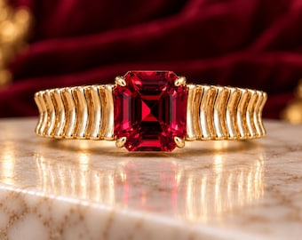 May include: A gold ring featuring a large, rectangular, emerald-cut red gemstone set in the center. The band is crafted from polished gold and displays a series of vertical, ribbed, or fluted ridges extending from the central setting.