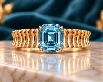 May include: A gold ring featuring a central emerald-cut light blue gemstone held by four prongs. The band is constructed from a series of vertical, curved gold bars that create a ribbed texture extending around the finger.