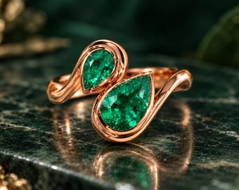 May include: A bypass ring crafted from rose gold, featuring two pear-cut green gemstones set in smooth, polished metal bezels. The design showcases a continuous, flowing band that curves around the fingers, highlighting the vibrant green stones.