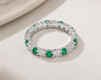 May include: A silver eternity band ring featuring an alternating pattern of round-cut clear white stones and round-cut vibrant green stones set in a prong setting. The inner band is clearly engraved with the hallmark S925, indicating sterling silver.