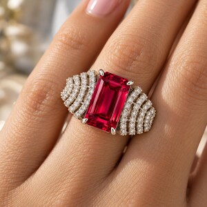 Peut inclure: Une bague argent&eacute;e orn&eacute;e d'une grande pierre pr&eacute;cieuse rectangulaire rouge vif taille &eacute;meraude, entour&eacute;e de chaque c&ocirc;t&eacute; par trois rang&eacute;es incurv&eacute;es de petites pierres claires rondes serties pav&eacute; sur un anneau m&eacute;tallique.