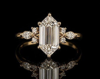 May include: A gold ring featuring a large, elongated emerald-cut center diamond held by six prongs. The band is accented on each side by a cluster of two small round diamonds and one marquise-cut diamond, all set in a polished yellow gold metal band.