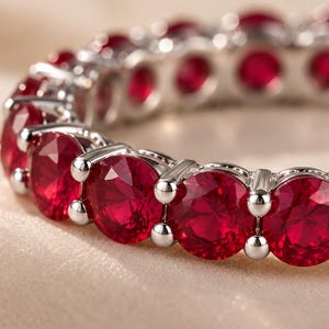 May include: An eternity band ring featuring a continuous row of round-cut, vibrant red gemstones set in a polished silver-toned metal setting. The circular design showcases multiple faceted stones held securely by small metal prongs in a row.
