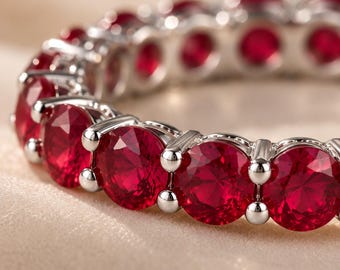 May include: An eternity band ring featuring a continuous row of round-cut, vibrant red gemstones set in a polished silver-toned metal setting. The circular design showcases multiple faceted stones held securely by small metal prongs in a row.