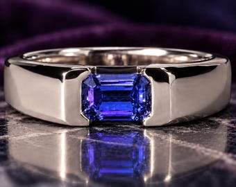May include: A polished silver-toned metal ring featuring a single rectangular emerald-cut blue gemstone set horizontally in the center. The band has a smooth, wide, and solid construction that frames the vibrant blue stone securely.