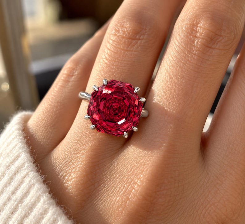 May include: A silver ring featuring a large, round, faceted deep red gemstone set with multiple prongs. The stone has a complex, rose-like geometric cut that reflects light across its surface, secured by a polished silver band on a finger.