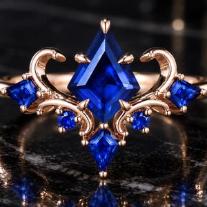 Op de afbeelding: Een roségouden ring met een grote, vliegervormige blauwe saffier als middelpunt, geaccentueerd door kleinere vliegervormige en ronde blauwe saffieren. De metalen band vertoont gedetailleerde krulmotieven die de centrale blauwe edelstenen omlijsten.
