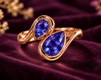 May include: A bypass ring crafted from polished rose gold, featuring two pear-shaped, deep blue faceted gemstones set in a smooth, curved metal frame that wraps around the finger with an open design.