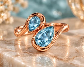 May include: A bypass ring crafted from rose-toned metal, featuring two pear-shaped, faceted light blue gemstones set in smooth bezels. The open-ended design showcases a polished, reflective finish that highlights the vibrant blue stones.