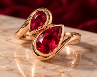 May include: A bypass ring crafted from polished rose gold, featuring two pear-cut, deep red rubies set in smooth bezels. The design showcases a fluid, open-ended band that curves around the finger, highlighting the vibrant, faceted gemstones.