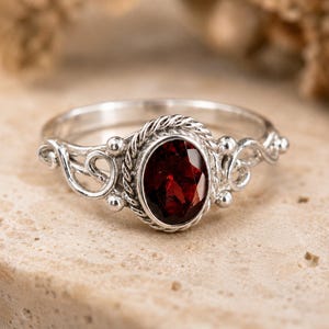 Könnte beinhalten: Ein Ring aus Sterlingsilber mit einem zentralen, oval geschliffenen, tiefroten Granat-Edelstein. Der Stein ist in eine gedrehte Kordelbordüre gefasst und wird von dekorativen Silberverzierungen sowie kleinen Kugelakzenten flankiert.