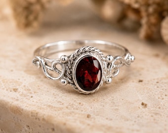 Peut inclure: Une bague en argent sterling ornée d'une pierre précieuse grenat rouge foncé de taille ovale. La pierre est sertie dans une bordure torsadée, flanquée de volutes en argent décoratives et de petits accents sphériques sur les côtés.