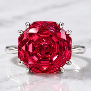 May include: A ring featuring a large, vibrant red gemstone with a multifaceted, geometric rose-like cut. The stone is secured by eight silver-toned prongs on a smooth, polished silver-toned metal band, creating a bold and symmetrical appearance.