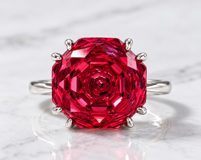 May include: A ring featuring a large, vibrant red gemstone with a multifaceted, geometric rose-like cut. The stone is secured by eight silver-toned prongs on a smooth, polished silver-toned metal band, creating a bold and symmetrical appearance.