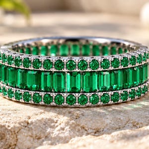 Peut inclure: Une alliance argentée composée d'une rangée centrale de pierres vertes taille émeraude, encadrée par deux rangées de pierres vertes plus petites taille ronde. La bague présente une monture métallique maintenant les pierres en cercle.
